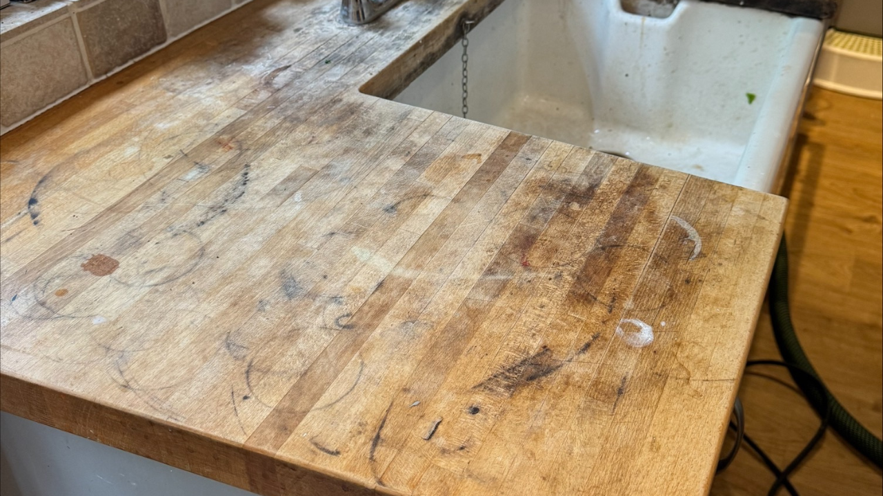 Worktop Refinishing in Gants Hill on a solid oak butcher-block kitchen worktop, shown heavily stained and worn beside a white sink with oak plank hardwood floor in the background.